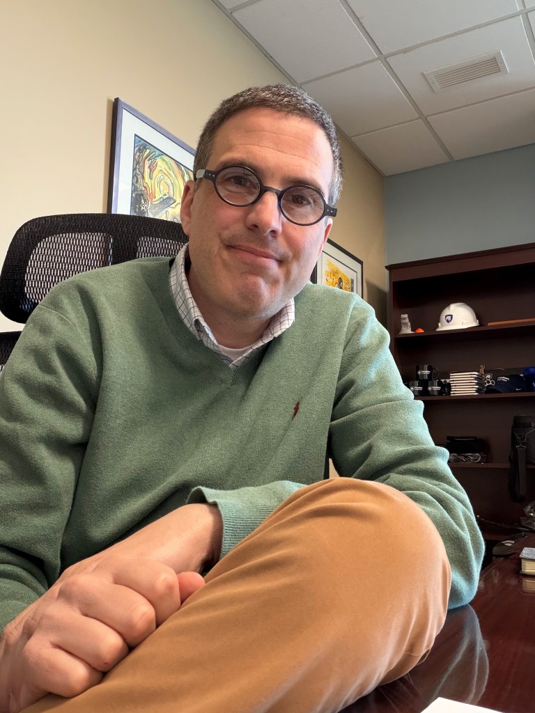 Ryan Abramson Director of Strategic Communications at Penn State Lehigh Valley sitting in his office at the Center Valley, PA campus with a bookshelf and Nittany Lion hat behind him.